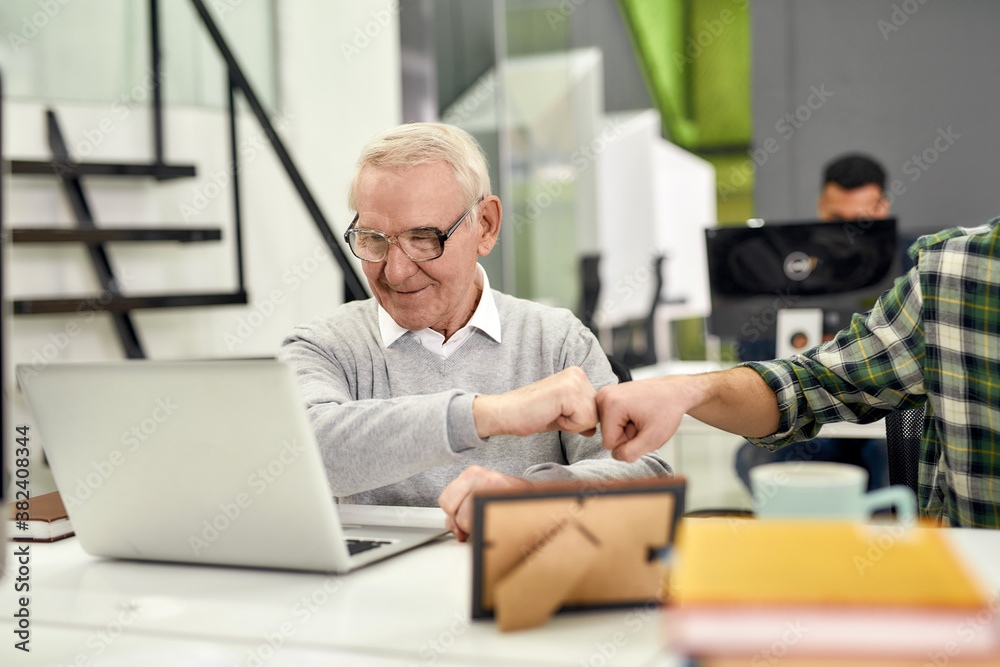Aged man, senior intern looking at the screen of his laptop and doing a fist bump with his young colleague, Friendly male worker engaging new employee while working in the office Aged man, senior intern looking at the screen of his laptop and doing a fist bump with his young colleague, Friendly male worker engaging new employee while working in the office
