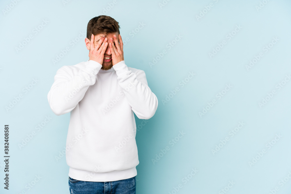 Young caucasian man isolated on blue background blink at the camera through fingers, embarrassed covering face. Young caucasian man isolated on blue background blink at the camera through fingers, embarrassed covering face.