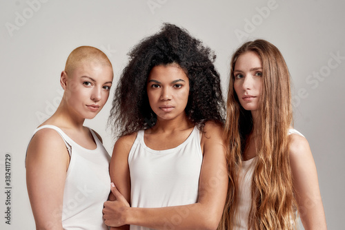 Portrait of three young diverse women wearing white shirts looking at camera while posing together isolated over grey background