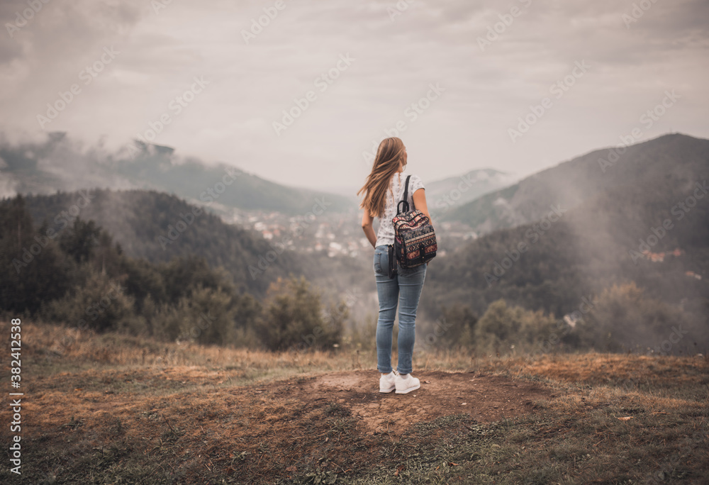 Young blonde woman standing alone with backpack on wild forest mountains at background. European girl wear tight jeans and white t-shirt sneackers. Carpathian Mountain peaks in fog scenery landscape