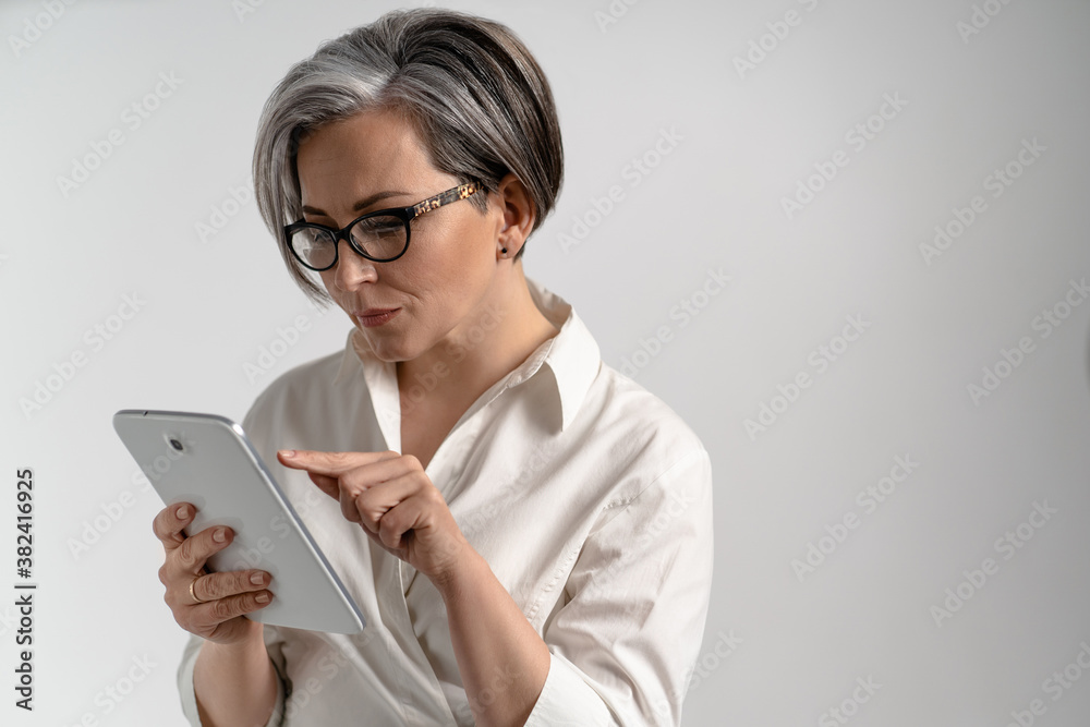 Elegant woman with glasses holds tablet and presses her finger. Mature woman with gray hair in white shirt on an isolated background. High quality photo.