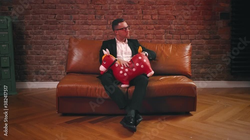 Portrait of young man in eyeglasses and suit sitting on leather coach spanking inflatable red horse