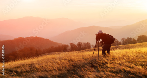 Photographer taking photo with tripod of autumn landscape with foggy peaks and orange trees. Ukrainian Carpathians mountains