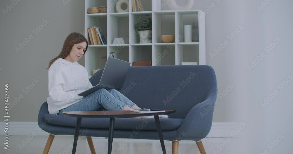 Low angle view of a woman sitting on a sofa looking at her laptop. Freelance Career. Cheerful Lady Working On Laptop Computer Sitting On Sofa At Home. Low angle view of a woman sitting on a sofa looking at her laptop. Freelance Career. Cheerful Lady Working On Laptop Computer Sitting On Sofa At Home.