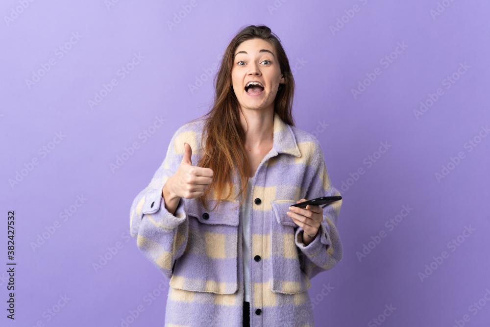 Young Ireland woman isolated on purple background using mobile phone while doing thumbs up Young Ireland woman isolated on purple background using mobile phone while doing thumbs up
