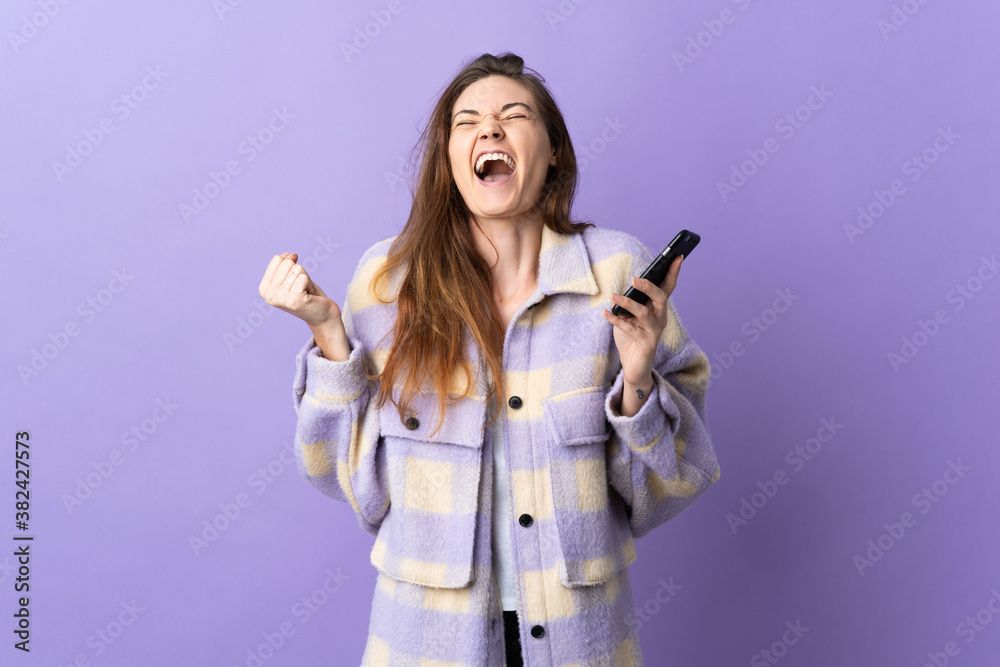 Young Ireland woman isolated on purple background using mobile phone and doing victory gesture Young Ireland woman isolated on purple background using mobile phone and doing victory gesture