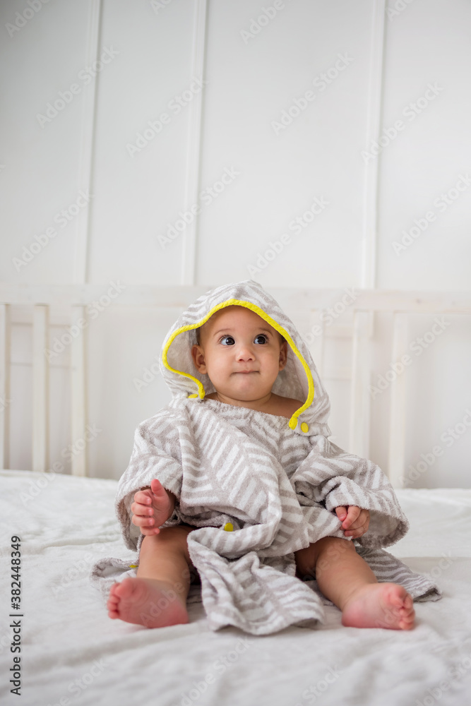 a serious little boy sits in a hooded bath towel on a white bed and looks away. Vertical orientation