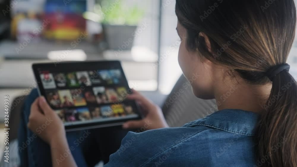 Caucasian woman lying down on sofa in living room at home searching for film or TV series to watch for leisure. Hispanic girl choosing movie on internet streaming service. Over the shoulder view