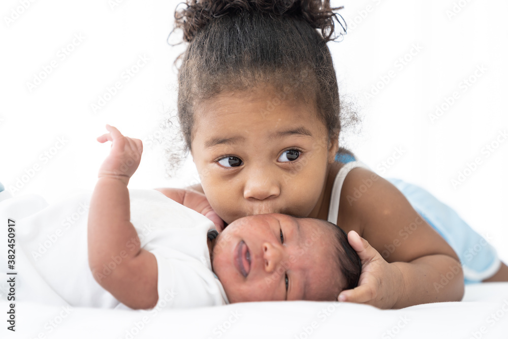 Cute African American little girl kissing on newborn baby cheek on white bed at home. Little girl takes care of infant baby with kindly Cute African American little girl kissing on newborn baby cheek on white bed at home. Little girl takes care of infant baby with kindly
