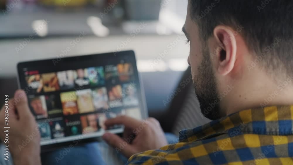 Young white man lying down on sofa in living room at home searching for on line films or TV series to watch for fun. Latino people choosing for movies on internet streaming service