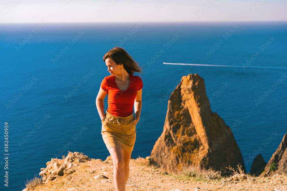 A beautiful young woman stands high above sea, on the background of coastal cliffs, calm clear blue sea, cape Fiolent in Balaklava Crimea.