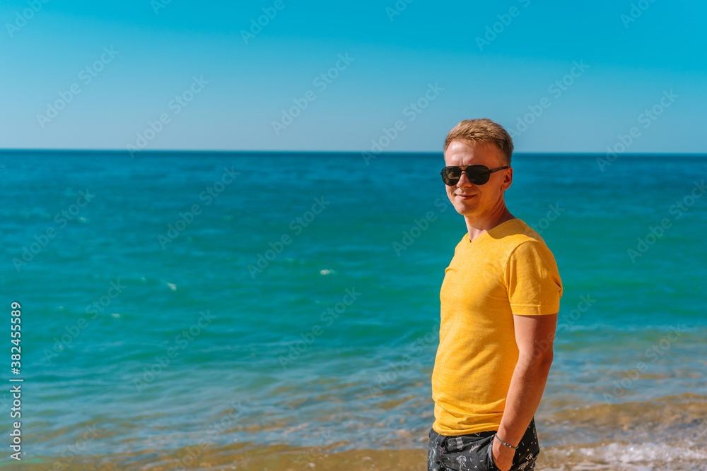 A young man in bright clothes and sunglasses walks on a sandy beach near the sea