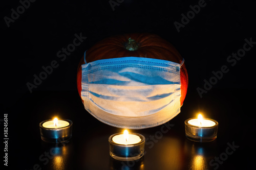 Pumpkin in medical disposable mask in dark and three lighted tea candles. Halloween and new normal concept