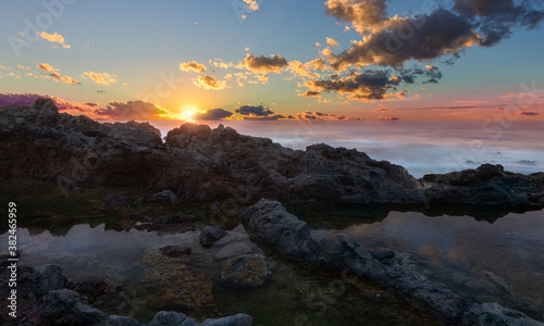 A romantic sunset on the rocky north coast of Tenerife. A natural pool is separated from the sea by rocks. The sky is reflected in the smooth water.