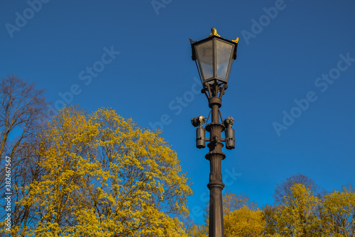 Old vintage black decorative lantern with glass lamps on pillar, pole in bright yellow spring forest. Blue sky. Saint Petersburg city park in sun light