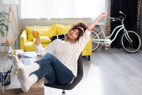 Slika na platnu curly woman with closed eyes holding glass of orange juice while resting on chai