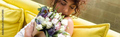 panoramic crop of young woman holding bouquet and smelling flowers in living room