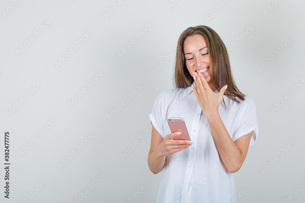 Young woman using smartphone with hand on mouth in white t-shirt and looking happy , front view. Young woman using smartphone with hand on mouth in white t-shirt and looking happy , front view.