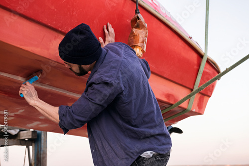 Bearded man in hat painting boat in red color