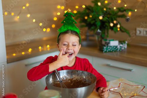Happy little girl prepares cookies on the eve of christmas. A child in the kitchen waiting for the new year. A girl in the kitchen surrounded by Christmas tree decorations and Christmas garlands.