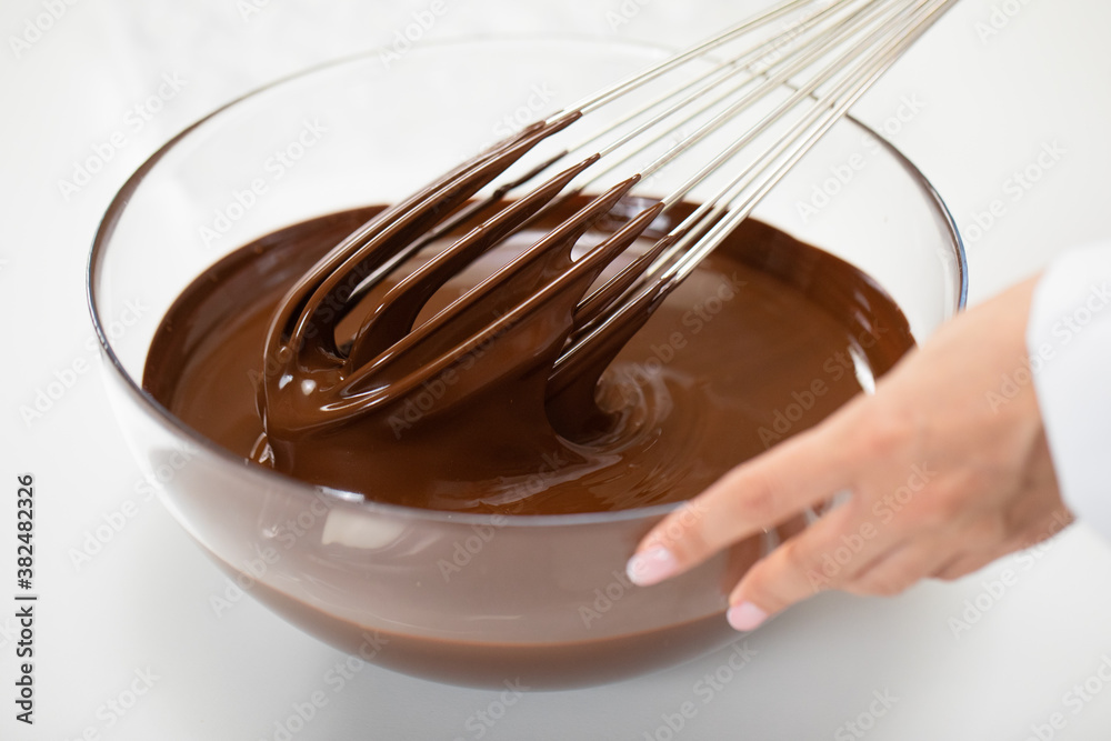 Fototapeta premium Hand of Female chocolatier mixing melted premium dark chocolate with whisk isolated on white close up. Melted Chocolate Swirls In glass Bowl. World Chocolate Day