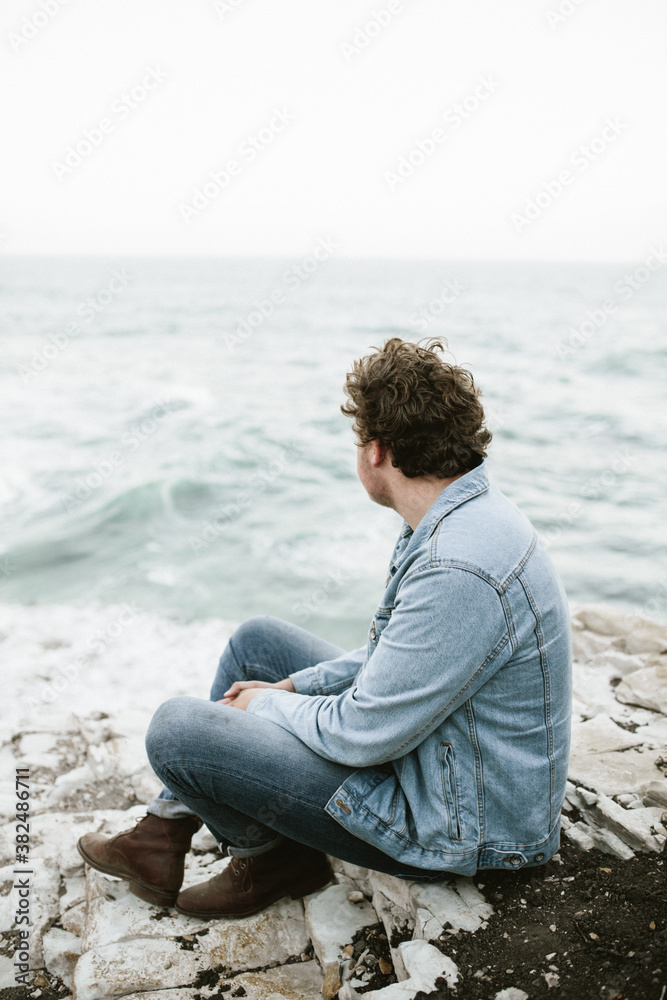 young man in denim jacket sitting by light blue ocean on white rocks young man in denim jacket sitting by light blue ocean on white rocks