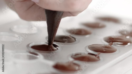 cropped view of chocolatier hand with pastry bag pouring melted dark chocolate into candy mold preparing luxurious handmade confectionery slow mo. World Chocolate Day