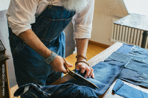 Closeup of Cool Senior Man Cutting Denim Fabric on Living Room Table photo