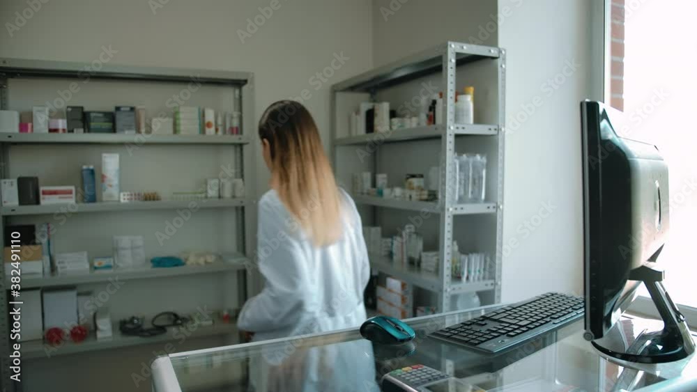 Caucasian woman checking order on computer and going to shelf with medicines. Daylight bright pharmacy interior. Computer display, keyboard, electronic mouse, glass showcase.