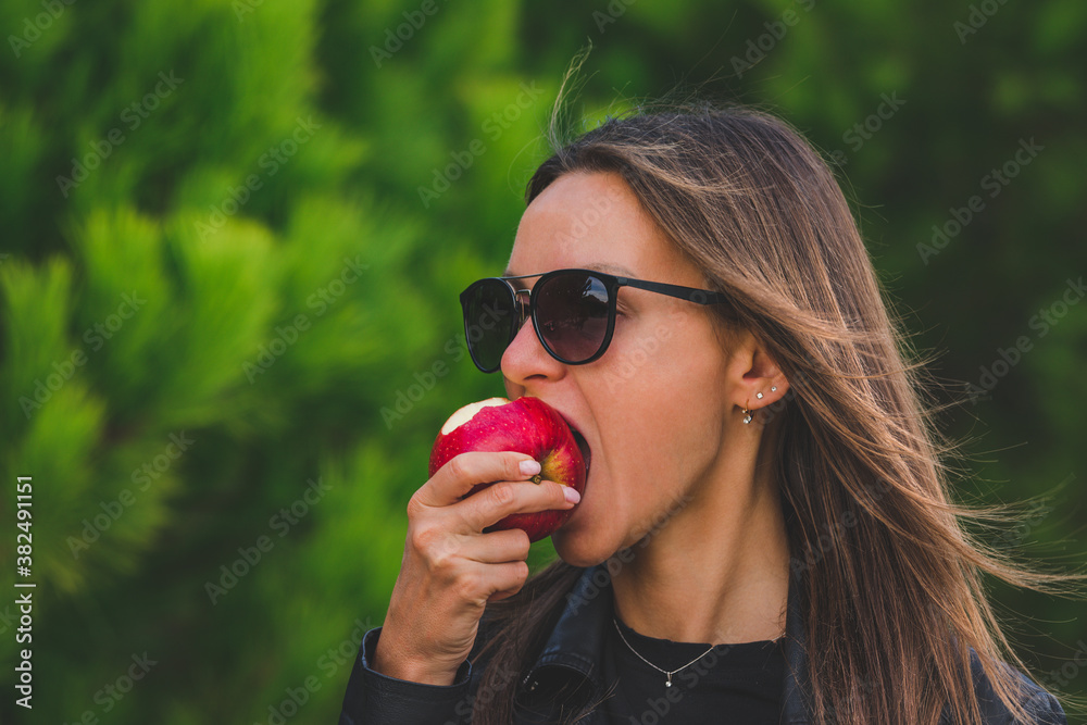 Beautiful young brunette vegan woman in black sunglasses eating red tasty apple on green trees background. Bite the apple. Healthy lifestyle concept. Beautiful young brunette vegan woman in black sunglasses eating red tasty apple on green trees background. Bite the apple. Healthy lifestyle concept.