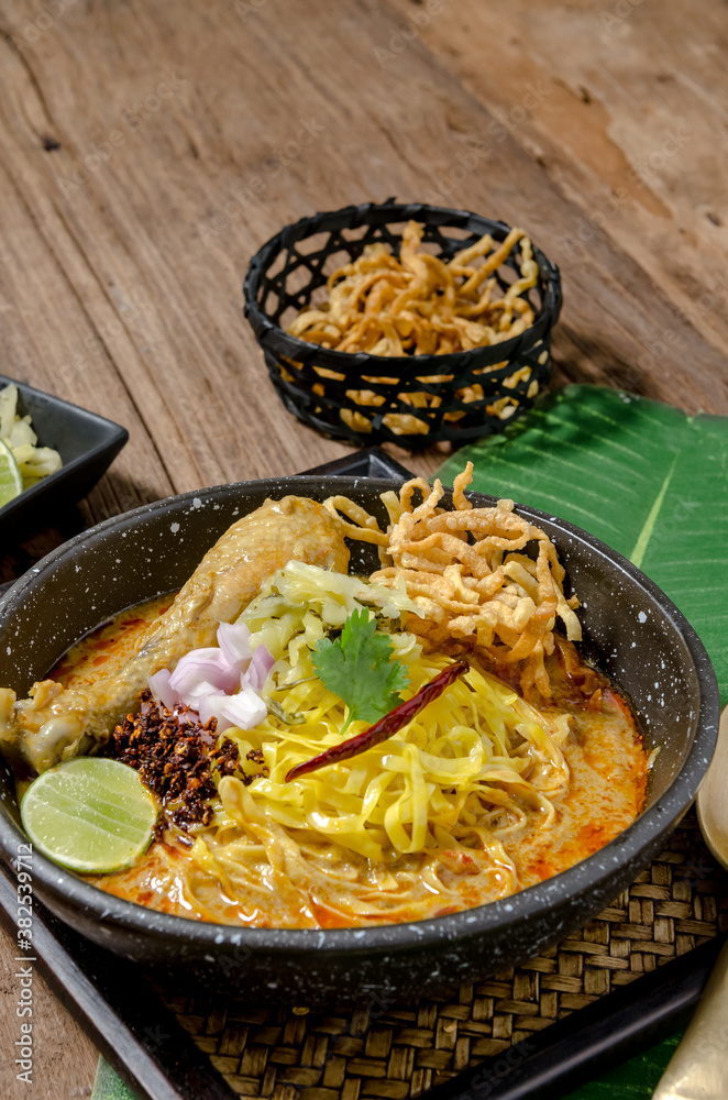 The tasty egg yellow noodle in chicken curry soup with ingredients in the bowl on wooden table. Kao Soi Kai, this is name of the traditional food of northern Thailand.. The tasty egg yellow noodle in chicken curry soup with ingredients in the bowl on wooden table. Kao Soi Kai, this is name of the traditional food of northern Thailand..