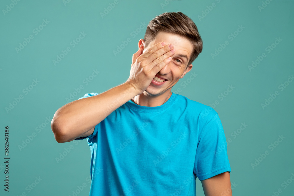 Happy male portrait. Peek-a-boo fun. Playful mood. Excited emotion. Young smiling man closing eye hand looking at camera isolated on blue. Happy male portrait. Peek-a-boo fun. Playful mood. Excited emotion. Young smiling man closing eye hand looking at camera isolated on blue.