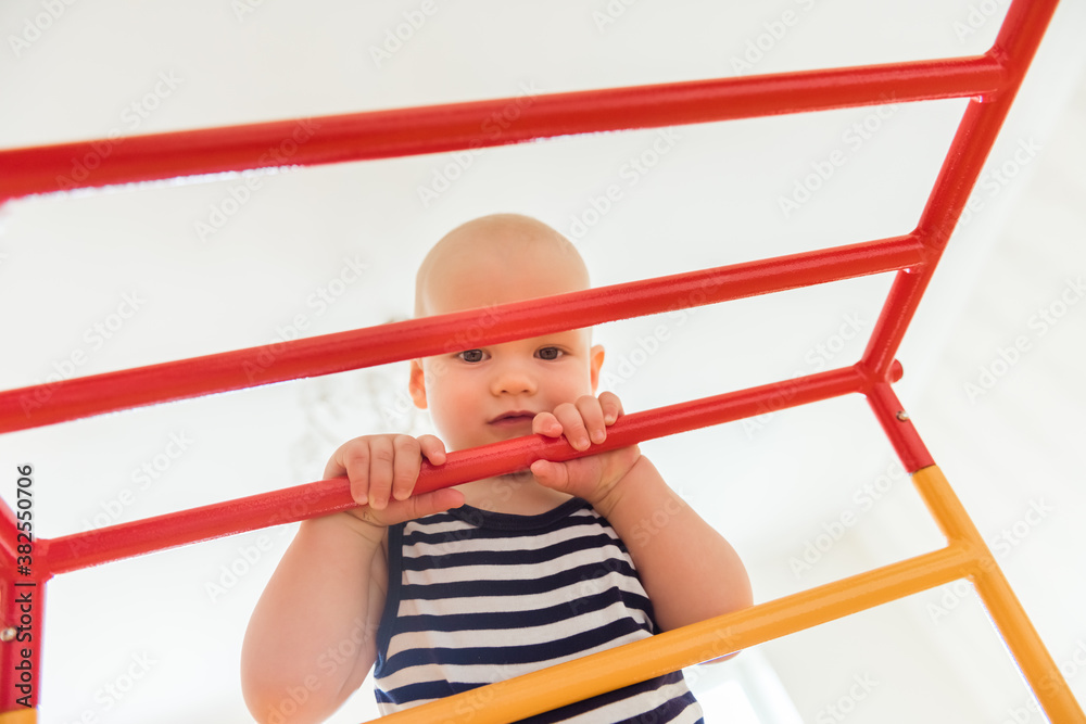 Cute baby performs gymnastic exercises on a wooden home sports complex stairs and rings. Children's sports exercises. Physical education of children at home Cute baby performs gymnastic exercises on a wooden home sports complex stairs and rings. Children's sports exercises. Physical education of children at home