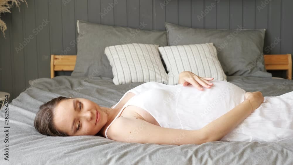 Happy pregnant woman in white dress lies on bed stroking tummy at home close-up Happy pregnant woman in white dress lies on bed stroking tummy at home close-up