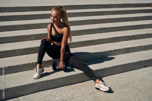 A young woman athlete trains biceps with a dumbbell on the steps. A blonde in a black suit is engaged at dawn. Healthy lifestyle.