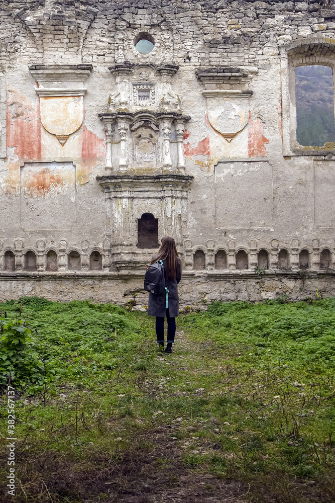 Autumn trip,  travel, vacation, weekend: young woman, tourist with a backpack and warm coat stands in dilapidated old synagogue. Back view. Rashkov, Moldova. Selective focus.