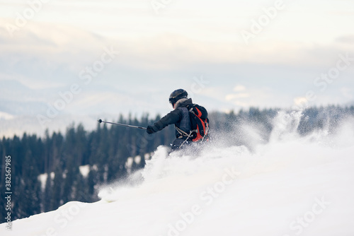 Back view of skier backpacker descending from mountain in deep white snow powder. Skier on high slope. Concept of popular winter extreme amateur sport. Mountains forest view. Grey sky on background.