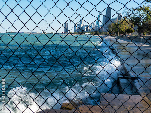 view through fence of waves spraying up on lakefront of Lake Michigan with view of downtown Chicago