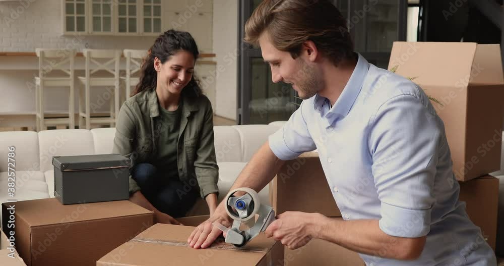 Happy married couple packing cardboard boxes with stuff preparing family belongings with adhesive tape while move-out from rented house at own new dwelling smile look at camera feels proud and excited