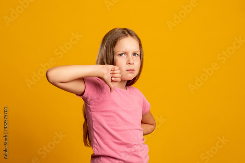 Dislike gesture. Disappointed child. Wrong choice. Bad idea. Sad young girl rejecting offer with thumb down looking at camera isolated on orange copy space background.