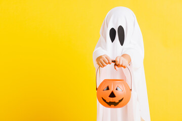 Funny Halloween Kid Concept, little cute child with white dressed costume halloween ghost scary he holding orange pumpkin ghost on hand, studio shot yellow on white background