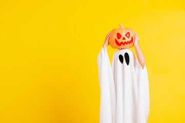 Funny Halloween Kid Concept, little cute child with white dressed costume halloween ghost scary he holding orange pumpkin ghost on hand, studio shot yellow on white background