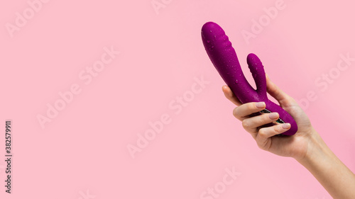 The girl's hand holds a purple vibrator covered with drops on a pink background. Vibrator for female masturbation. Dildo for vaginal and clitoris stimulation. Image for a sex shop. Sex toys for adult