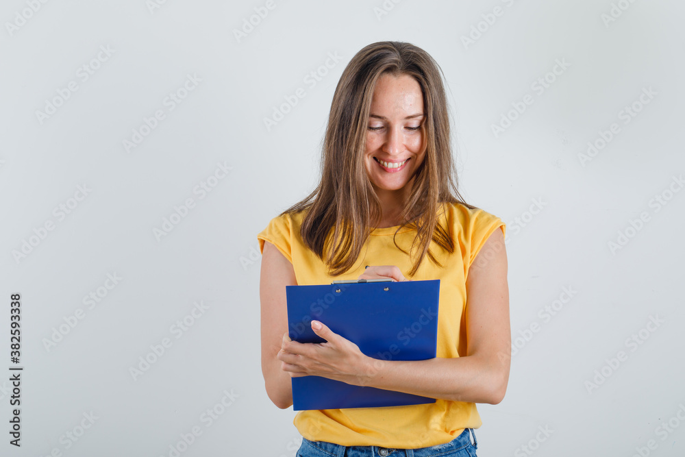 Young woman taking notes on clipboard in t-shirt, shorts and looking busy , front view. Young woman taking notes on clipboard in t-shirt, shorts and looking busy , front view.