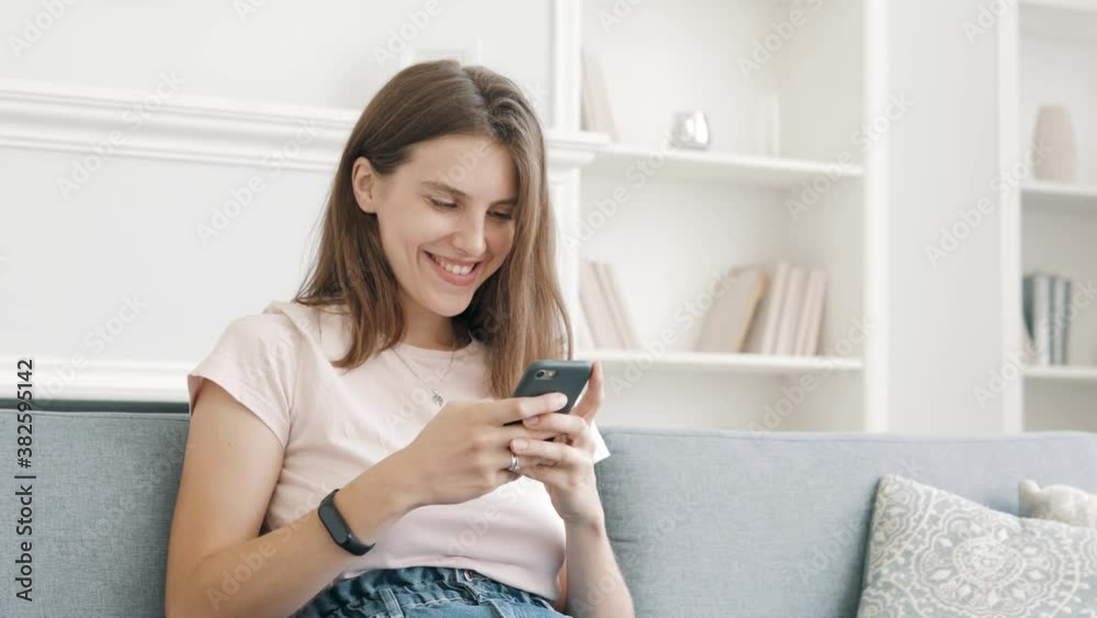 Happy relaxed young woman holding smart phone looking at cellphone screen laughing enjoying using mobile apps for shopping having fun playing games chatting in social media sit on couch at home Happy relaxed young woman holding smart phone looking at cellphone screen laughing enjoying using mobile apps for shopping having fun playing games chatting in social media sit on couch at home