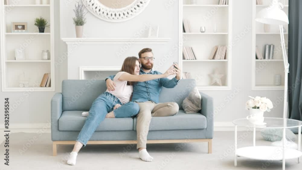 Carefree young adult couple enjoying relaxing using modern smartphone technology. Man and woman making video call playing mobile games, watching video, taking selfie sitting on a sofa together at home Carefree young adult couple enjoying relaxing using modern smartphone technology. Man and woman making video call playing mobile games, watching video, taking selfie sitting on a sofa together at home