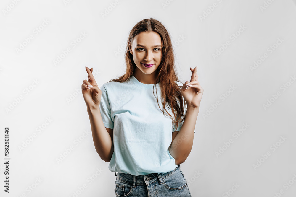 Beautiful young girl smiles crossed her fingers for luck and made a wish on a white isolated gray background. Place for advertising. Positive woman with fashion make-up with purple lips. Beautiful young girl smiles crossed her fingers for luck and made a wish on a white isolated gray background. Place for advertising. Positive woman with fashion make-up with purple lips.