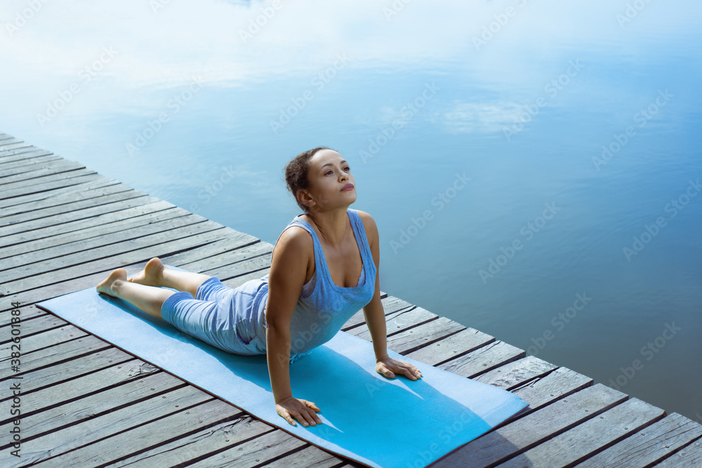Young woman on the dock doing asanas on the yoga mat and smiling. Outdoor yoga classes. Young woman on the dock doing asanas on the yoga mat and smiling. Outdoor yoga classes.