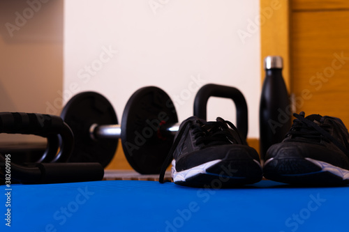 Ground level photography of sports equipment, dumbbells, sneakers, kettlebell, water bottle and push-up accessory, all on a mat for sports at home