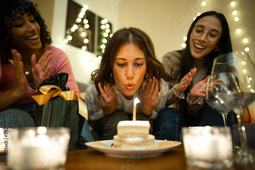 Close up of cute young woman blows on the cake candle next to her present celebrating her birthday at home with her two mixed race hispanic and caucasian female best friends